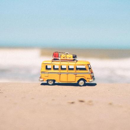 A vibrant yellow toy van sits on a sandy beach with an ocean backdrop, symbolizing fun travel adventures.