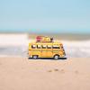A vibrant yellow toy van sits on a sandy beach with an ocean backdrop, symbolizing fun travel adventures.
