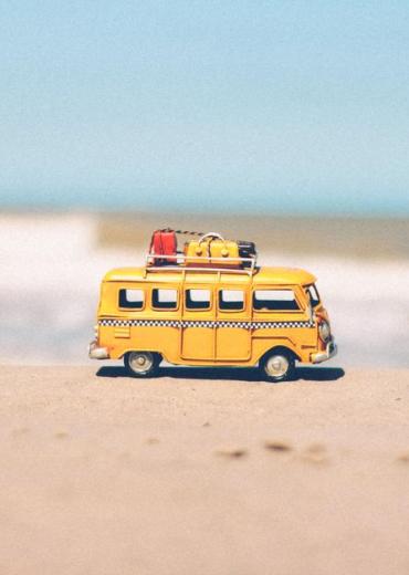 A vibrant yellow toy van sits on a sandy beach with an ocean backdrop, symbolizing fun travel adventures.